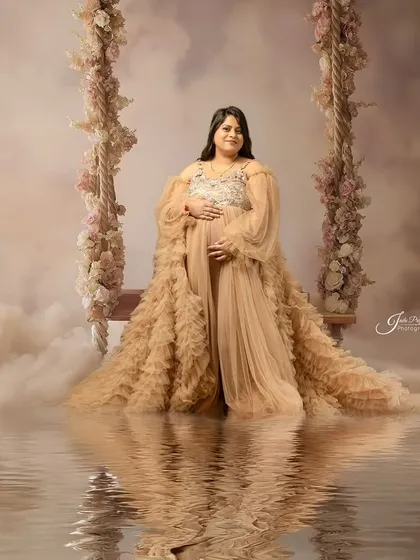 A dreamy and romantic fantasy portrait. The expecting mother stands on a swing surrounded by clouds and a water reflection, creating a beautiful, ethereal scene.