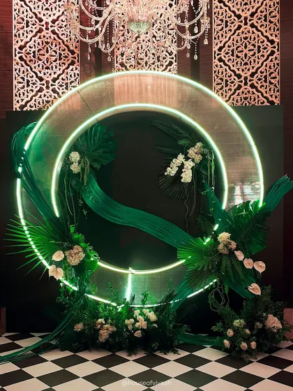 A bold and modern photo backdrop for a cocktail night, featuring a green neon halo, sculptural green fabric, and arrangements of tropical leaves and white anthuriums.
