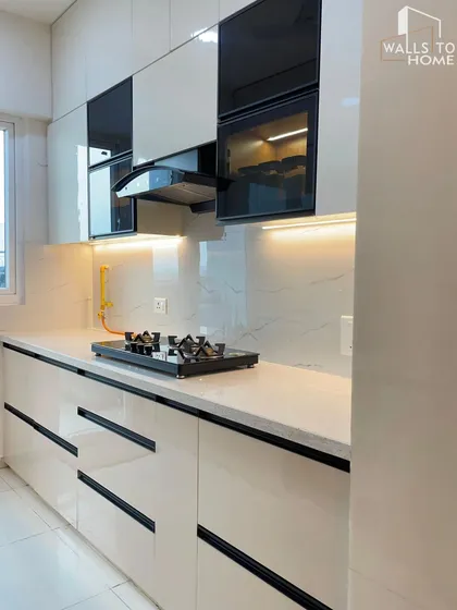 A modern kitchen with white high-gloss cabinets, black profile handles, and a built-in chimney. The under-cabinet lighting illuminates the white marble-patterned backsplash, creating a bright and clean workspace.