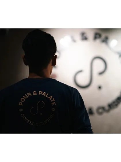 A shot from behind a barista at Pour & Palate cafe, with the cafe's logo softly focused in the background. It's a subtle branding shot that captures the atmosphere of the place.