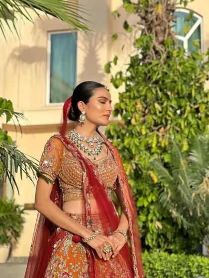 A bride enjoying a sun-kissed moment, her orange and red lehenga perfectly complemented by our elegant green and white Polki necklace set.