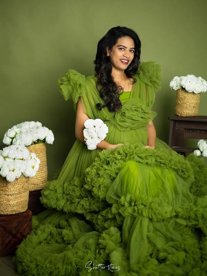 Seated amidst baskets of white flowers, the mom-to-be looks radiant in her green gown, creating a portrait that symbolizes life and growth.