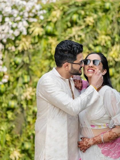A candid moment from a Haldi ceremony, where the couple shares a playful and affectionate look against a backdrop of white flowers and greenery.