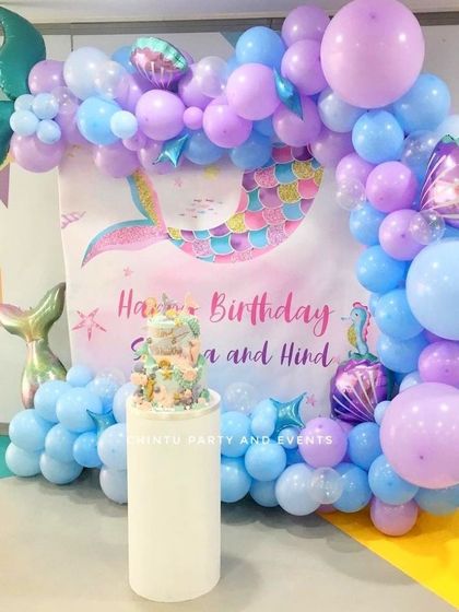 A magical mermaid theme birthday decoration using pastel blue and purple balloons. The setup includes a mermaid tail balloon and a themed backdrop for the cake table.