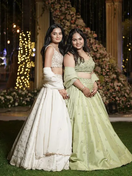 Two sisters pose together in their beautiful lehengas, one in ivory and one in mint green, creating a picture of coordinated elegance.