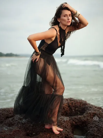 A dramatic beach portrait featuring a model in a black bodysuit and a sheer black tulle skirt. The pose is strong and the wind catches the skirt, creating a sense of movement against the ocean backdrop.