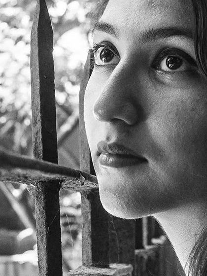 A close-up portrait of a woman looking through a barred window. Her expression is full of longing and story.