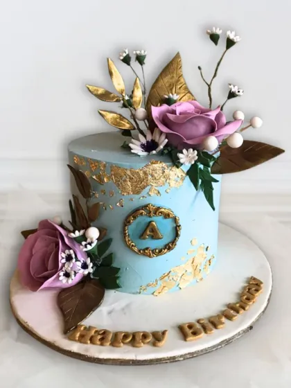 A beautiful light blue cake with gold leaf accents, a custom monogram, and an arrangement of delicate sugar flowers.