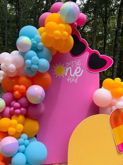 A bright and fun summer-themed birthday party. The decor includes a vibrant balloon garland and props like sunglasses and an ice cream pop.