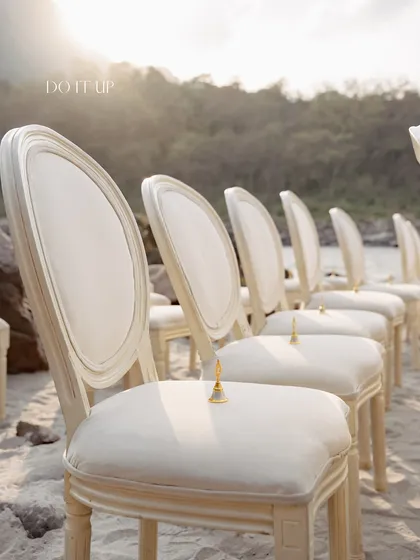 Simple elegance for a ceremony by the Ganges. Classic white chairs are lined up on the sand, each with a small golden bell for guests to ring, creating a serene and beautiful aisle.