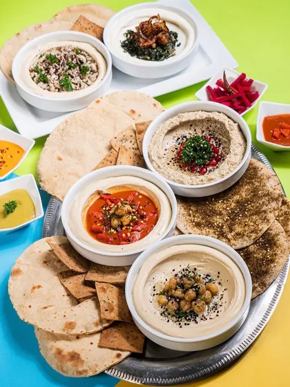 A platter of various hummus flavors and fresh pita bread, shot from above. The colorful background and arrangement of the dips create a visually appealing image that showcases the variety offered.