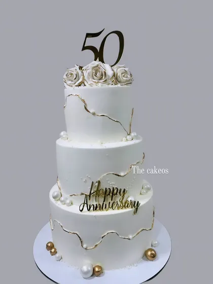 A stunning three-tier eggless cake created to celebrate a golden 50th anniversary. The design features elegant white fondant with delicate gold kintsugi-style cracks, pearls, and handcrafted sugar roses, perfect for a landmark celebration.