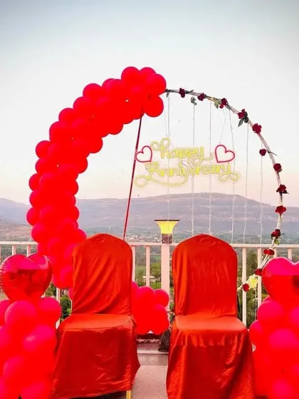 A romantic anniversary setup on a balcony with a scenic view. The decoration includes a red balloon arch, covered chairs, and a "Happy Anniversary" neon sign.
