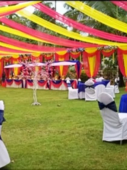 An outdoor wedding reception setup on a lawn with a canopy of yellow and pink drapes and guest tables dressed in blue and white.