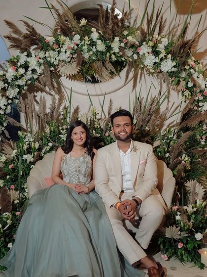 The couple at their boho-chic ring ceremony, seated on a shell-shaped sofa surrounded by a beautiful arch of pampas grass and white flowers.