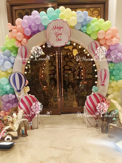 An enchanting hot air balloon themed entrance. A circular arch made of pastel rainbow balloons is decorated with hot air balloon cutouts and fairy lights, creating a magical entryway.
