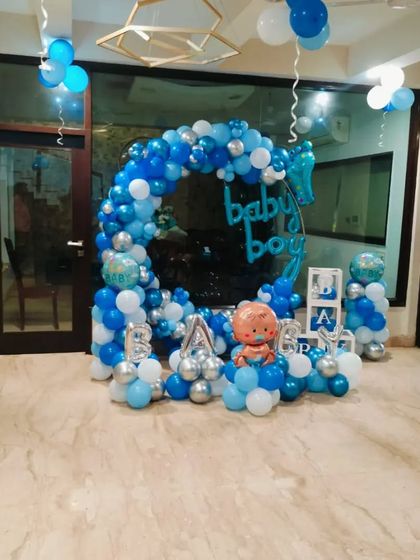 A full view of a "Baby Boy" welcome decoration with a circular balloon arch in blue, white, and silver. The setup includes "BABY" letter balloons and a cute baby boy foil character.