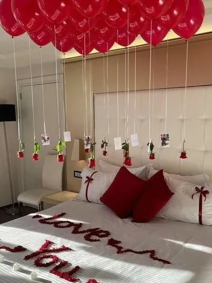 An elegant room decor idea for an anniversary or Valentine's Day. The setup includes red balloons with hanging photos and roses over the bed, with 'I Love You' written in petals.