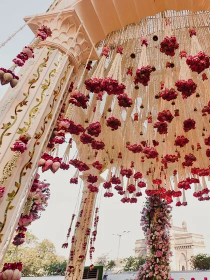 A close-up of the mandap ceiling, adorned with a dense canopy of red roses and lotus buds, with the Gateway of India visible in the distance.