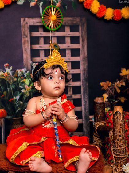 A beautiful portrait of a baby dressed as Krishna in a striking red outfit. The thoughtful upward gaze and traditional props make this a captivating image.