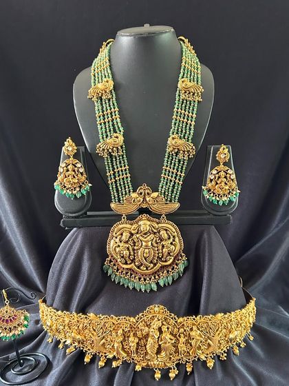 This antique finish bridal set features beautiful green beadwork and intricate deity carvings. The set includes a long haaram, matching earrings, maang tikka, and a detailed vaddanam.