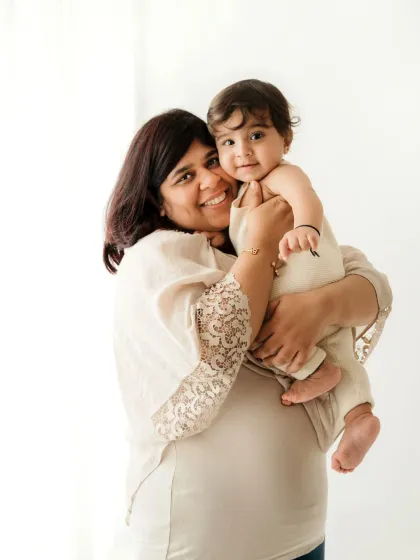 A mother holds her baby, both smiling warmly. The simple, bright background keeps the focus on their happy connection.