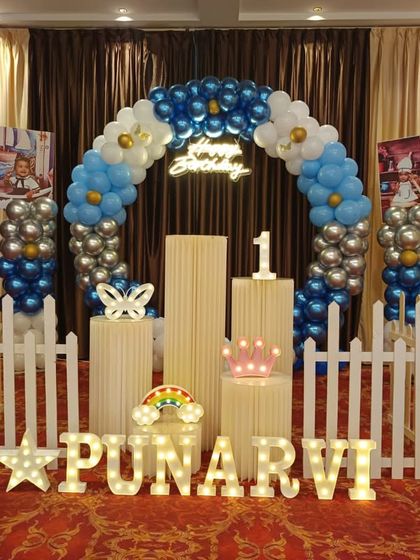 A blue and white themed first birthday decoration set up in a banquet hall. The arrangement includes a balloon arch, a white picket fence, and personalized marquee letters.