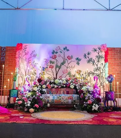 A wide view of a vibrant and artistic Mehendi stage setup. I used a custom-painted floral backdrop, a colorful patchwork sofa, and lush flower arrangements to create a festive and photo-ready space for the bride and guests.