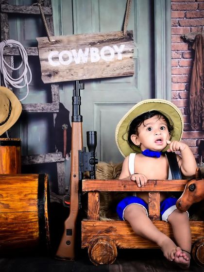 A little cowboy in a rustic wild west setup, complete with a hat, a toy rifle, and a wooden cart.