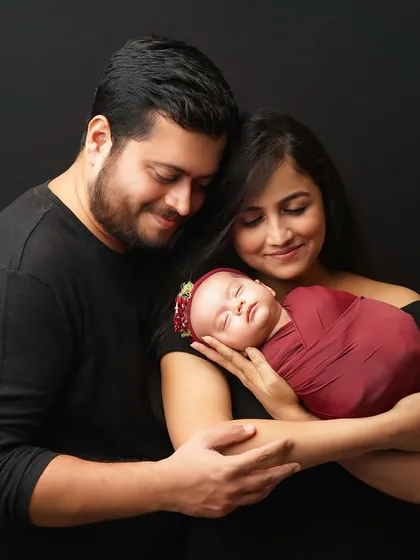 A classic family portrait against a simple black background. The new parents are smiling as they gaze down at their sleeping newborn, who is swaddled in a rich maroon wrap with a matching floral headband.