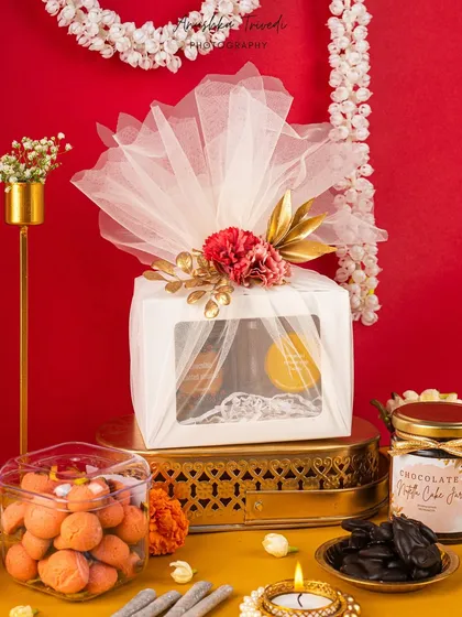 An elegantly wrapped white gift box, perfect for a premium Diwali hamper. The tulle wrapping, carnation, and golden leaves add a touch of sophistication and class.