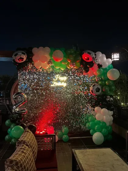 A stylish football-themed birthday party on a rooftop. This setup features a sparkling shimmer wall, a custom balloon garland, and a "Happy Birthday" neon sign, perfect for evening celebrations.