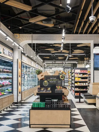 The interior of Le Marche streamlines the vibrant chaos of a traditional market into an ordered shopping experience. The checkerboard flooring and organized aisles guide customers, while displays like the fresh guacamole station add interactive elements.