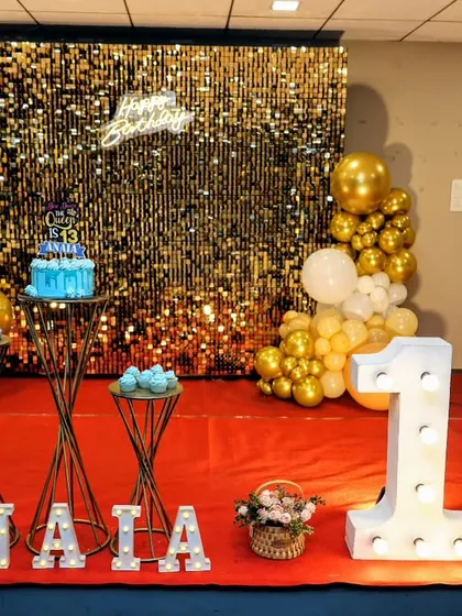 A glamorous 13th birthday party setup with a gold shimmer wall, a 'Happy Birthday' neon sign, and large marquee numbers. The gold and white balloons complete the luxurious look.