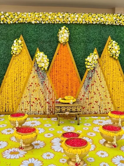 A unique geometric backdrop for a Haldi ceremony made with marigold flowers. The triangular panels and traditional 'kalash' props create a very distinct and modern look.