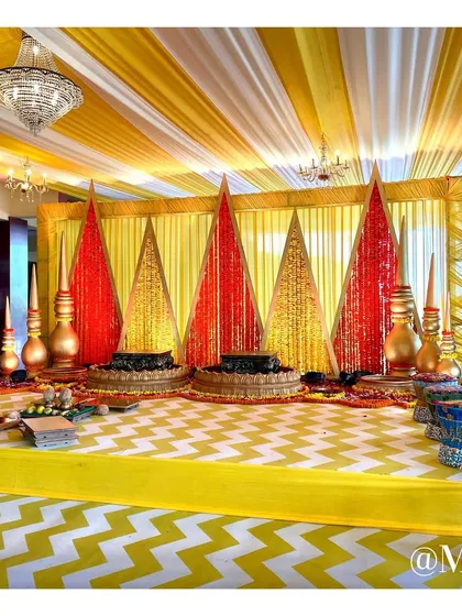 The main seating area for the Haldi ritual. The stage features lotus-shaped seats for the couple, set against a backdrop of triangular panels decorated with red and yellow flowers.