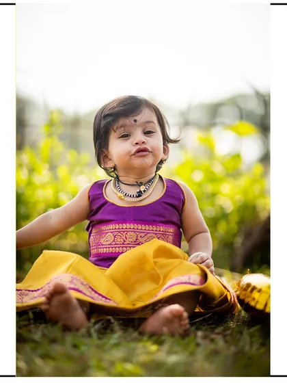 A vertical portrait of a baby girl in a traditional paithani-style dress, sitting happily in the sunlit grass.