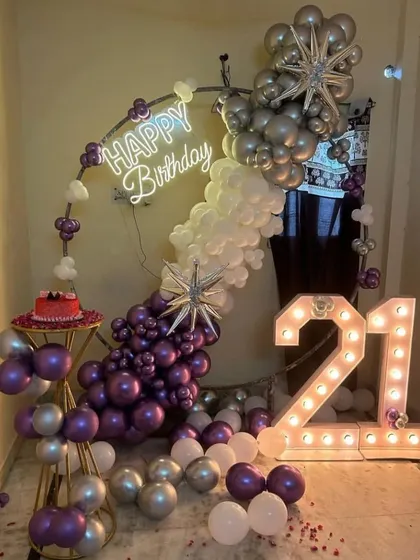 A glamorous 21st birthday celebration corner. This setup combines a circular balloon arch in purple, silver, and white with a "Happy Birthday" neon sign and large, illuminated "21" marquee numbers for a perfect photo opportunity.