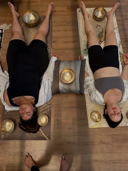 Two clients experiencing a semi-private sound healing session. Lying in 'Savasana' or the corpse pose, they are open to receiving the therapeutic frequencies, with bowls placed at their feet and head.