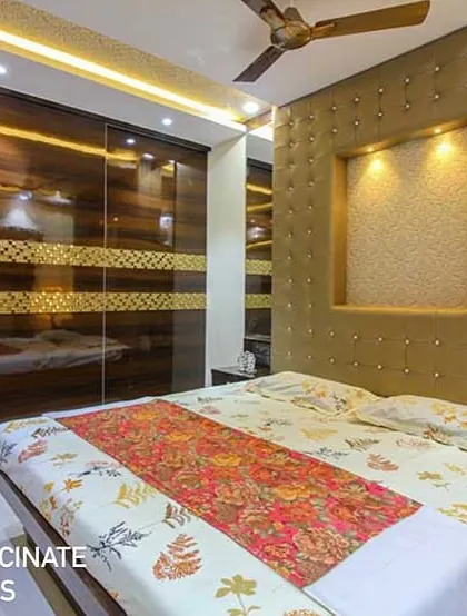 A bedroom in a Vashi row house designed with a rich, golden-upholstered headboard that includes a lit niche. The glossy, wood-grain wardrobe with a custom gold-patterned band adds a touch of opulence and warmth.