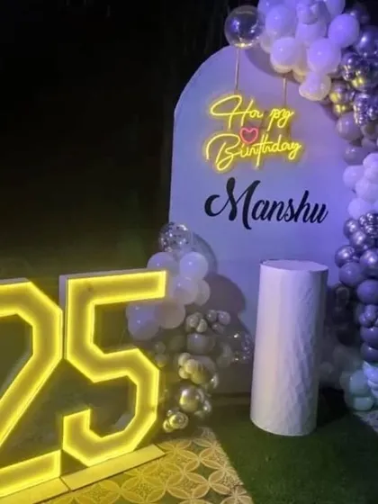 An outdoor 25th birthday party at night. This setup features a white arched backdrop, a purple and silver balloon garland, a neon sign, and large marquee numbers.