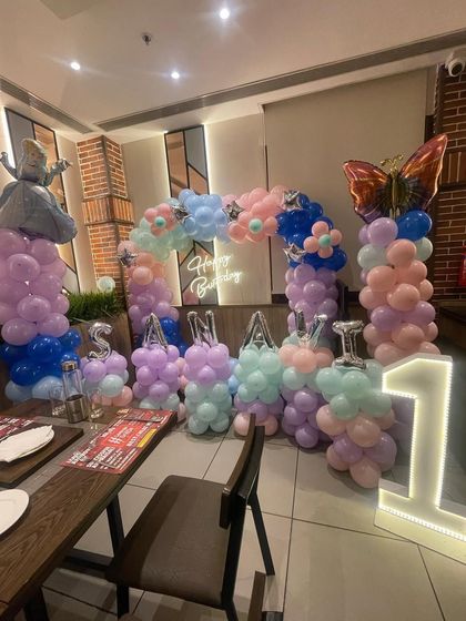 A beautiful pastel-colored decoration for a first birthday. This setup features a princess theme with an Alice in Wonderland foil balloon, a butterfly balloon, and marquee letters spelling 'SANAVI'.