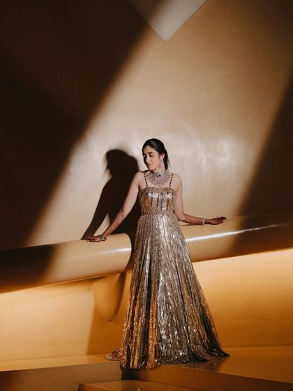 An elegant full-length portrait of the bride. The dramatic shadows and warm lighting create a cinematic feel, highlighting her poise and the shimmering details of her golden gown.