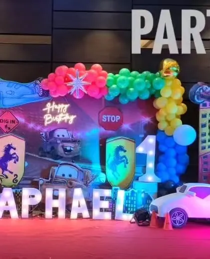 A close-up of a Cars themed birthday decoration, showing character cutouts, a light-up number '1', and marquee letters spelling "Raphael".