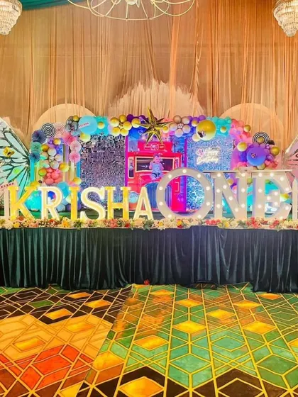 A spectacular butterfly-themed stage for 'KRISHA's first birthday, with large glittering wings, a sequin backdrop, and colourful lighting.
