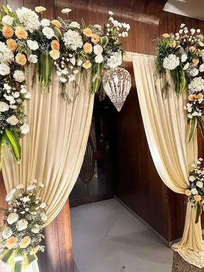 An elegant doorway treatment with an arch of white and peach flowers, soft cream-colored drapes, and a hanging crystal lantern. This classic design creates a graceful and welcoming entrance.