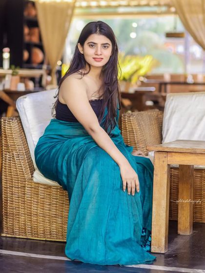 A modern take on saree styling. The model wears a teal saree paired with a contemporary black top, shot in a cafe with earthy tones. This shows how traditional wear can be styled for a modern, chic look.
