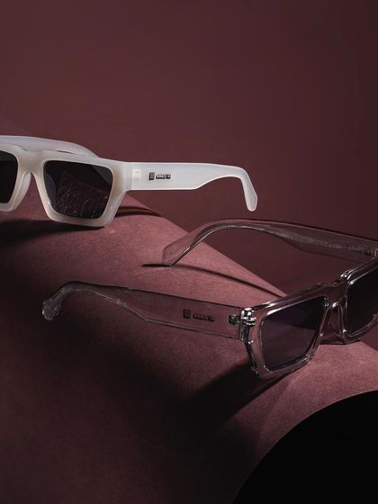 This sleek product shot showcases two pairs of sunglasses on curved, textured paper. The clean composition and sophisticated lighting highlight the modern design and different finishes of the eyewear.