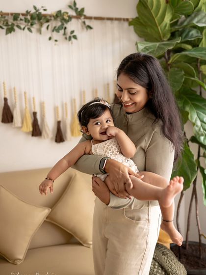 A mother holds her giggling baby girl in a cozy, sunlit corner of my studio. The baby's contagious smile is the highlight of this warm and loving portrait.