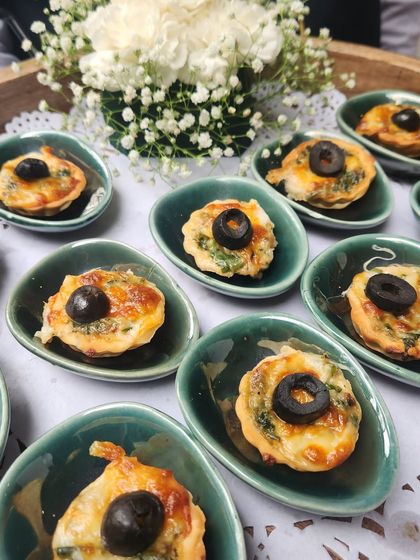 Golden, flaky Spinach and Corn Tarts topped with a black olive. These bite-sized quiches are served in beautiful ceramic dishes, adding a touch of elegance.